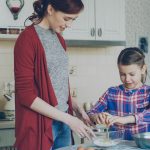 Little cheerful girl helping her mother in the kitchen cropping egg into bowl and mixing dough for cookies. Family, food, home and people concept