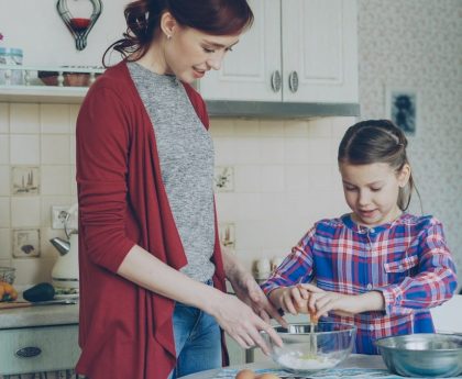 Little cheerful girl helping her mother in the kitchen cropping egg into bowl and mixing dough for cookies. Family, food, home and people concept